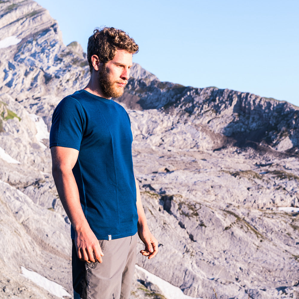 Ein Mann mit Bart in blauem Hemd steht vor einer Bergkulisse, passend zum Outdoor-Sortiment von alpinloakor.shop.