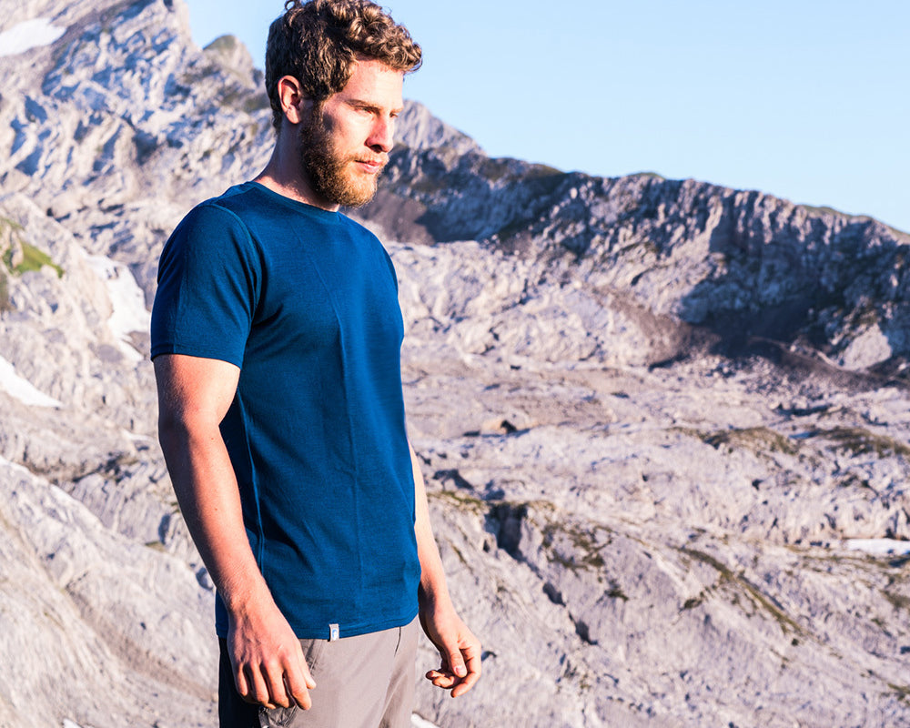 Ein Mann mit Bart in blauem Hemd steht vor einer Bergkulisse, passend zum Outdoor-Sortiment von alpinloakor.shop.