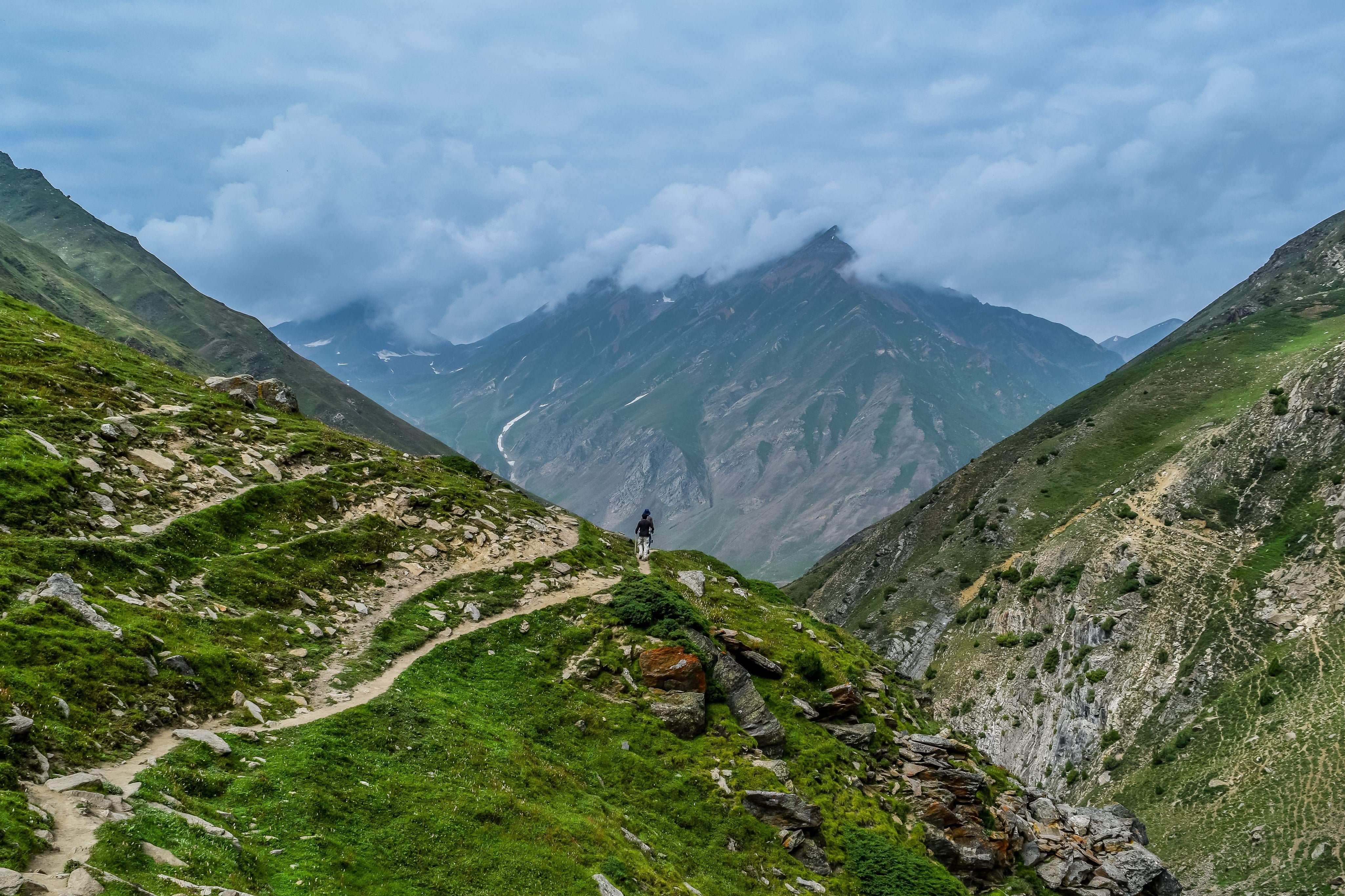 Eine Person wandert auf einem Bergpfad mit grünen Felsen und Gras, umgeben von einer Wolkenlandschaft.
