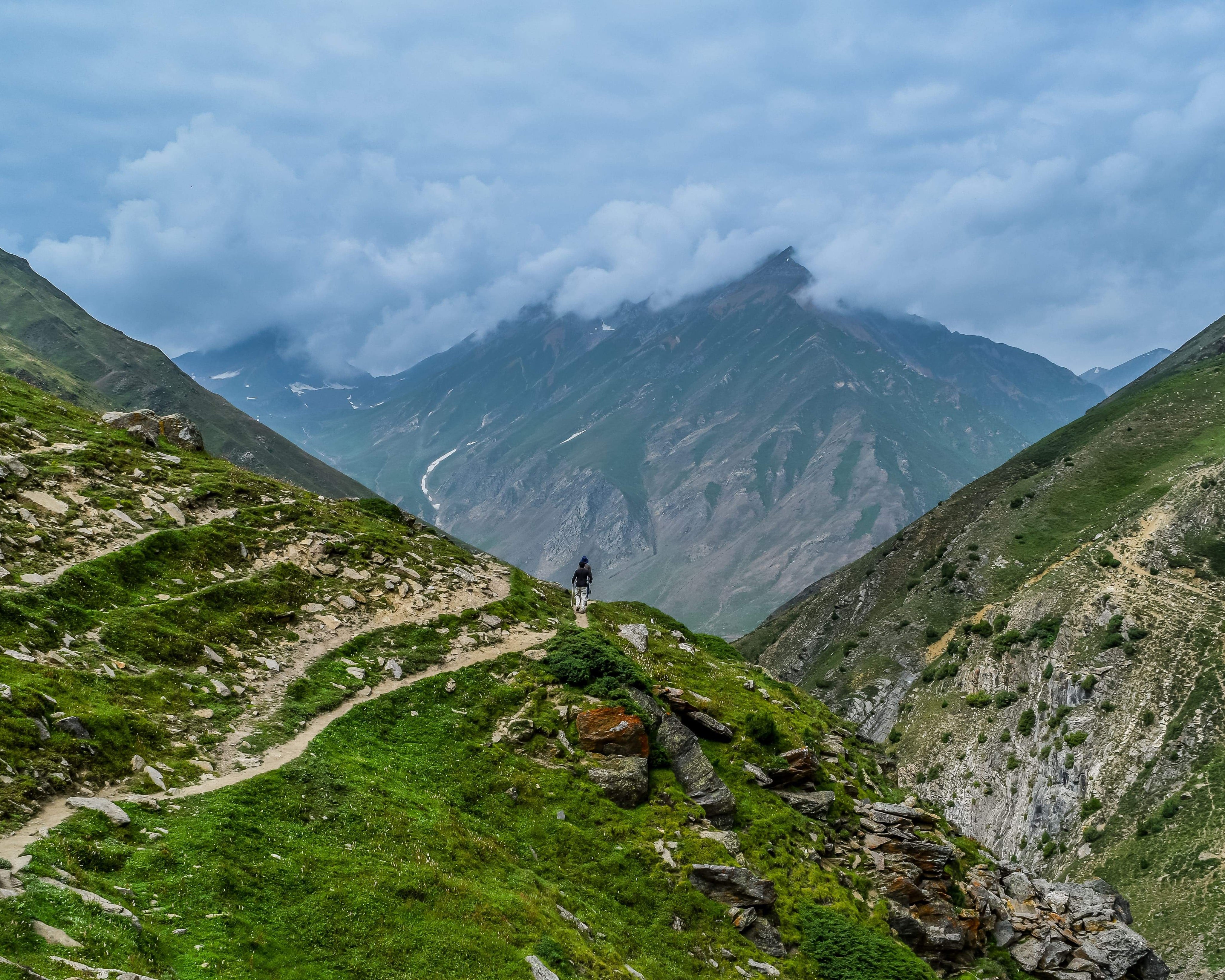 Eine Person wandert auf einem Bergpfad mit grünen Felsen und Gras, umgeben von einer Wolkenlandschaft.