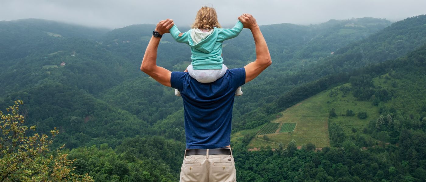 Ein Mann trägt ein Kind auf den Schultern beim Wandern, umgeben von Natur, passend zu Outdoor-Ausrüstung von alpinloakor.shop.