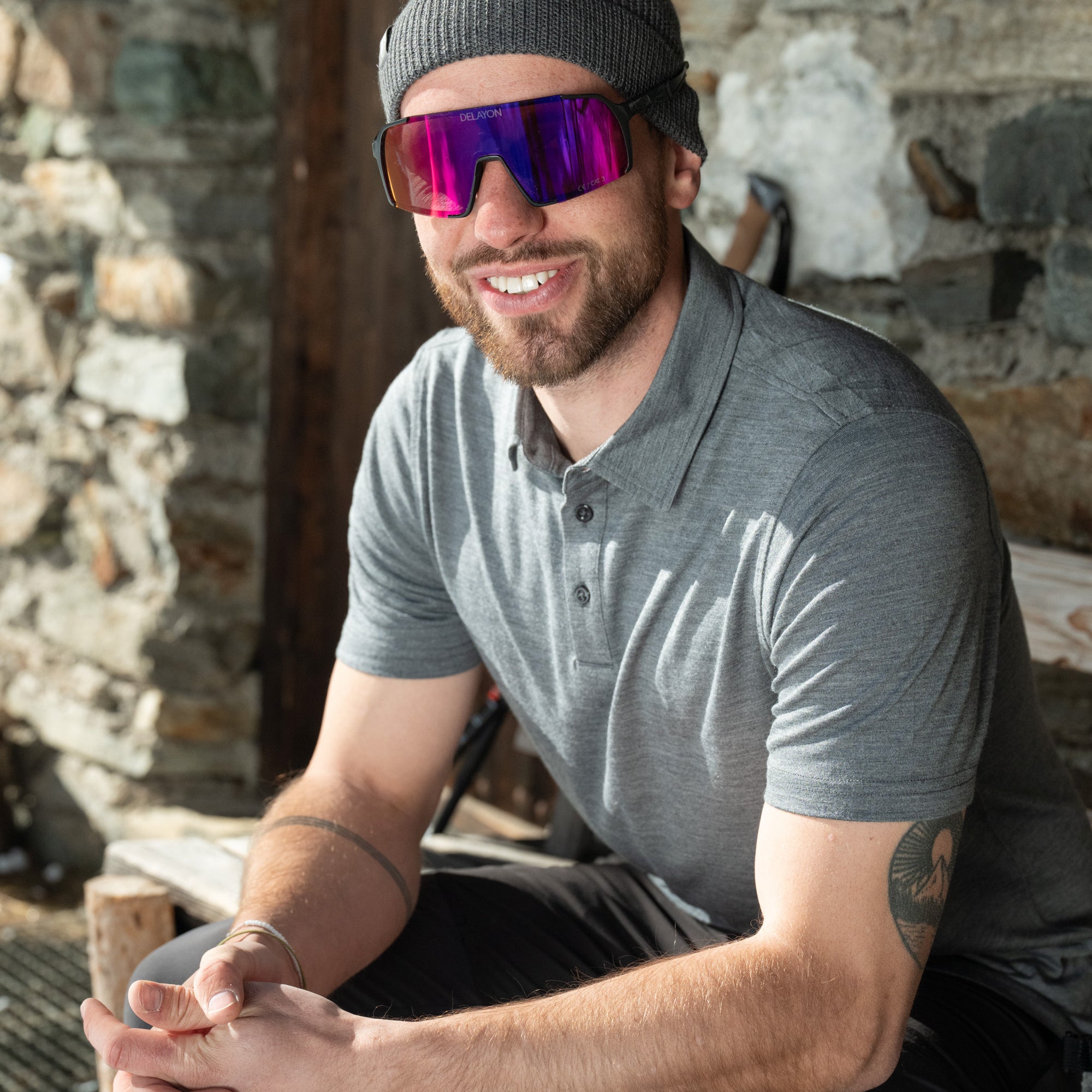 Model sitzt vor Hütte in grauem Merino Coolmax Polo Shirt - perfekt für Outdoor Aktivitäten