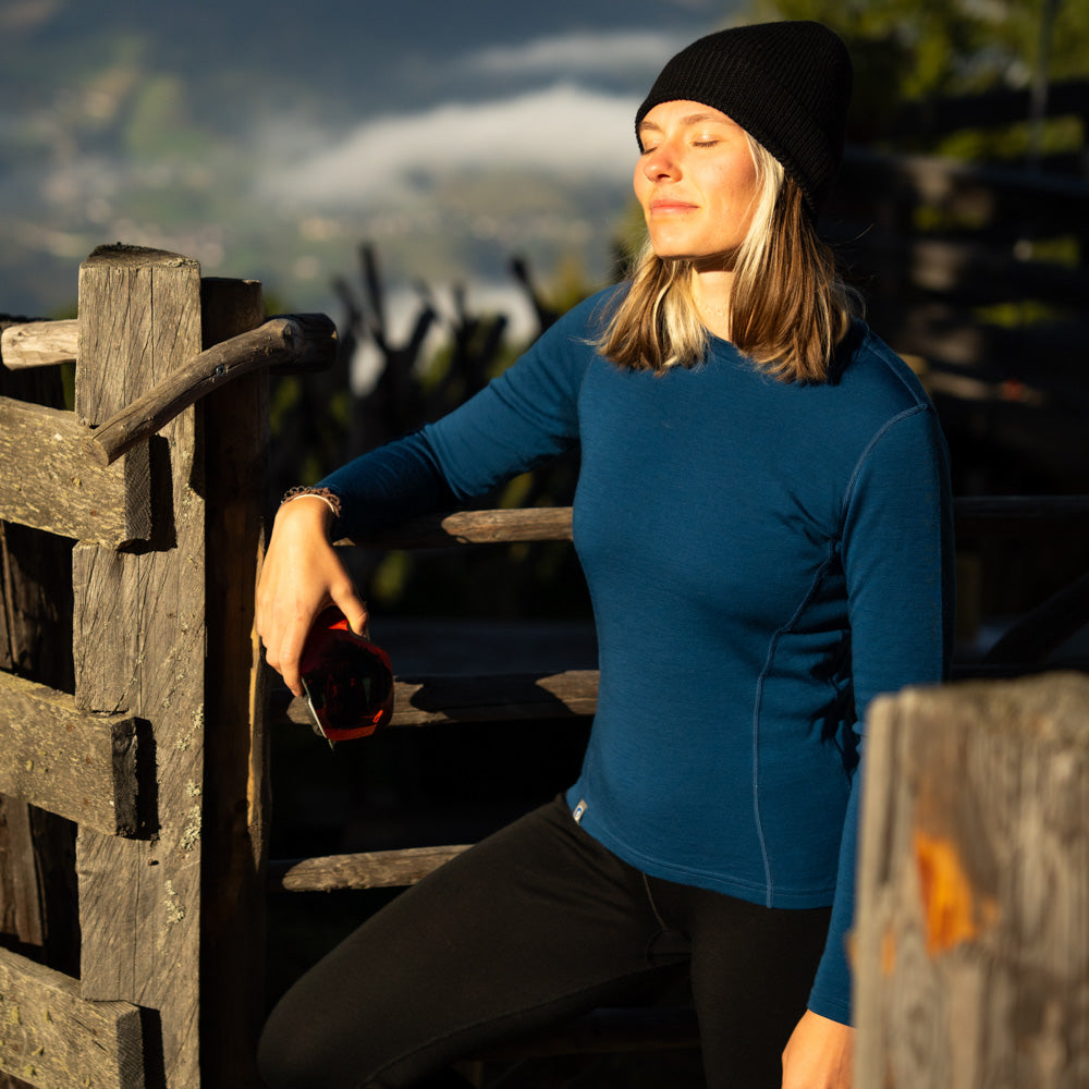 Eine Dame steht am Zaun in ihrem 230g Merino Set von Alpin Loacker. Das Oberteil ist blau. 