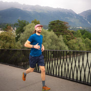 Mann joggt in Merino Coolmax T-Shirt Herren 150 g/m2 auf Brücke vor Bergkulisse, ideal für Outdoor-Aktivitäten durch temperaturregulierende und geruchsneutrale Eigenschaften.