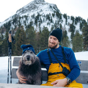 Mann mit Merino Mütze Herren & Damen Strick 230g/m² sitzt mit Hund und Stöcken vor schneebedecktem Berg, betont Wärme und Vielseitigkeit.