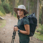 Junge Frau mit Sonnenhut mit UV Schutz Unisex auf Waldweg, bereit zum Wandern, trägt Trekkingstöcke und Smartwatch, verkörpert Abenteuergeist von alpinloakor.shop.