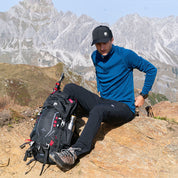 Ein Mann trägt ein Merino Langarmshirt mit Zip Herren 230 g/m², sitzt mit Rucksack auf Felsen in bergiger Landschaft. Ideal für Outdoor-Aktivitäten.