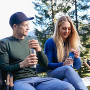 Zwei Wanderer, einer trägt das Merino Langarm Shirt Damen 230 g/m2, machen eine Pause im Wald mit Wanderstöcken und Wasserflaschen.