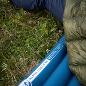 Ultra Leichte Isomatte für 3 Jahreszeiten auf Wiese mit Blumen, teilweise von grünem Schlafsack bedeckt, ideal für Trekking und Camping.