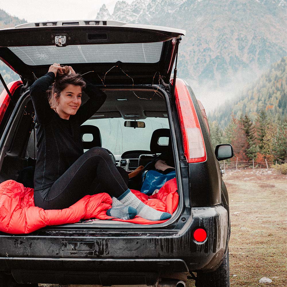 Frau im SUV mit Merino Langarm Shirt Damen 230 g/m2, ideal für Outdoor-Abenteuer, im Bergsetting mit Campingausrüstung und Lichterkette im Hintergrund.